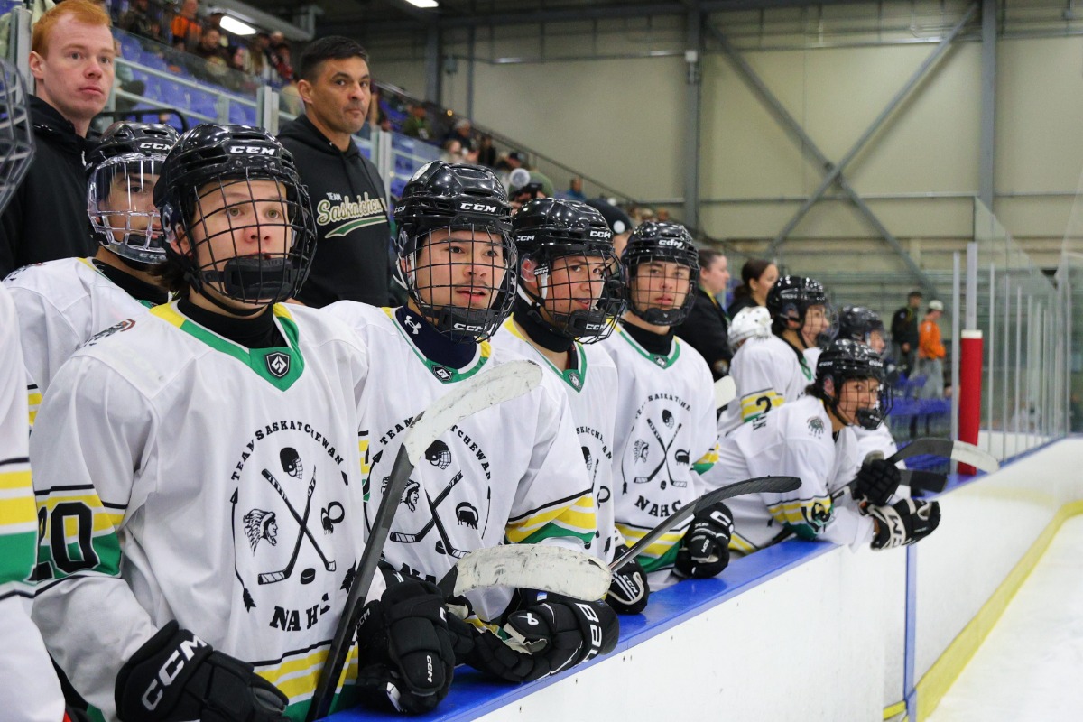 A young hockey team sits on the bench during a game. They are wearing Saskatchewan jerseys. They are all looking down the ice at something that's happening outside of frame.