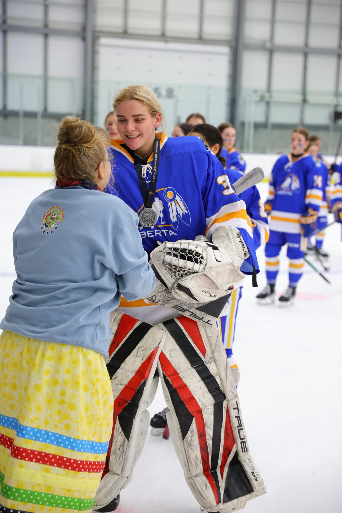 Team Alberta's goalie receives a medal from a woman in a yellow ribbon skirt.